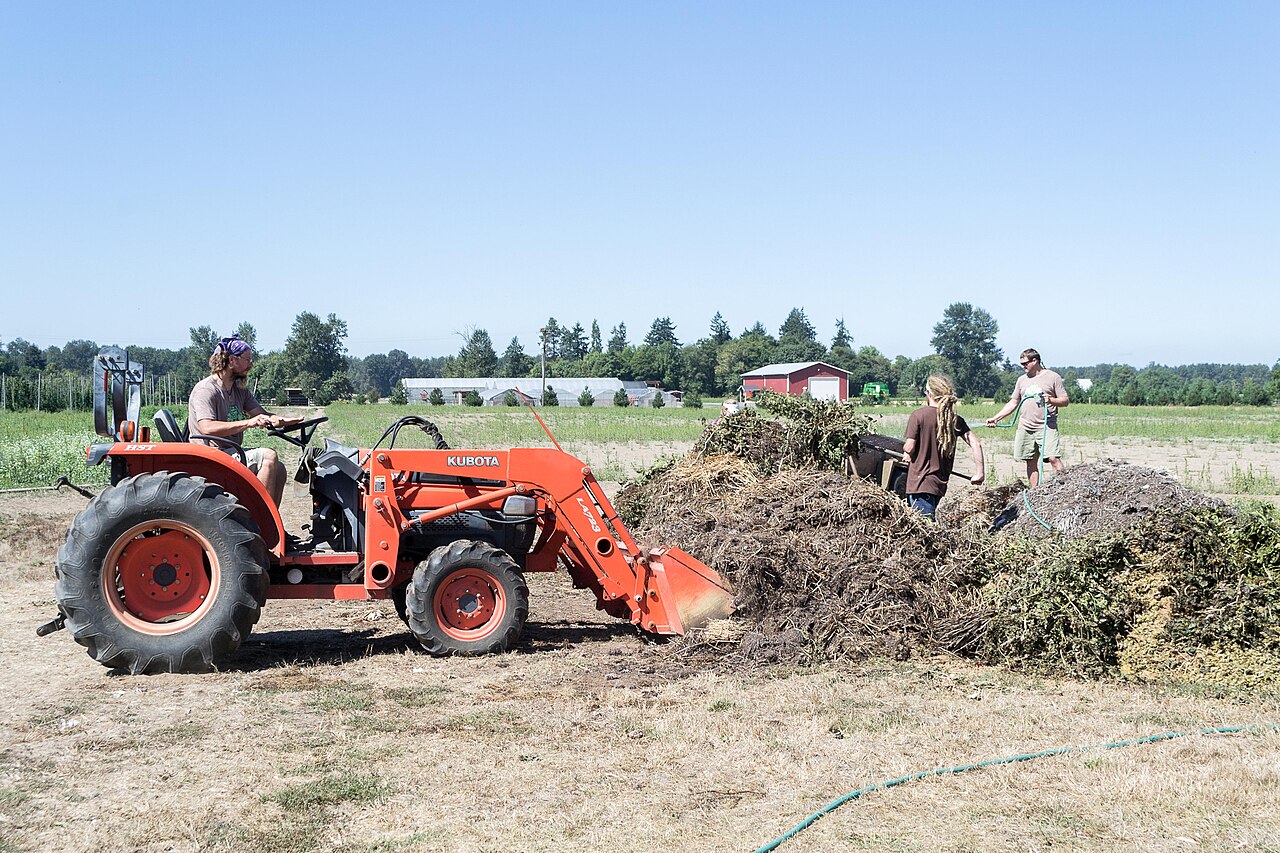 Volunteers and a tractor working beside large outdoor compost piles at an Oregon farm
