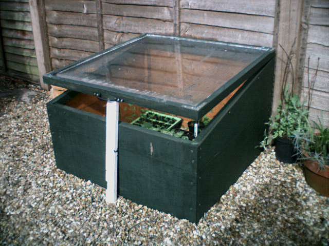 A cold frame with its lid propped open beside a garden fence